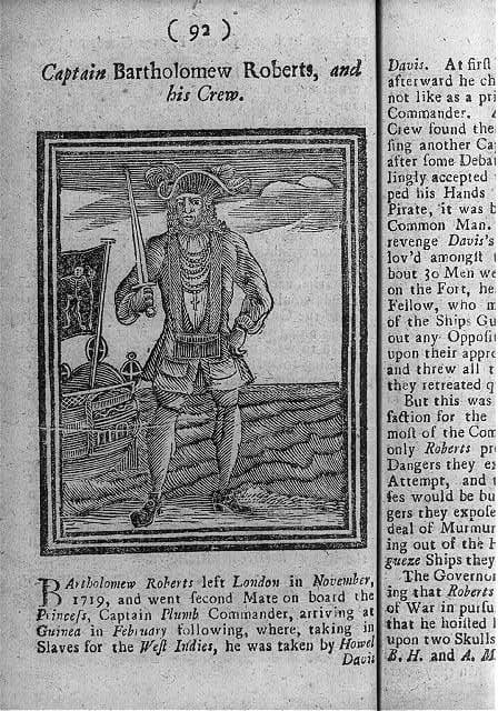A 1725 engraving of the pirate Bartholomew Roberts standing on a shore in a long coat and hat with a sword at his side and pistols slung across his chest, two ships lying at anchor behind him.